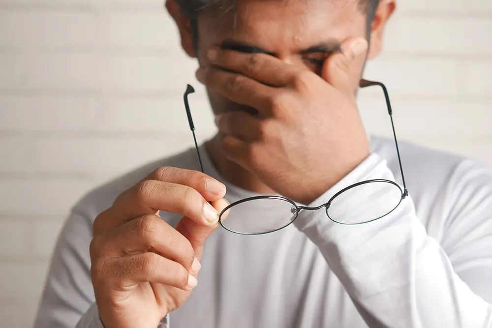 Man holding his prescription glasses in one hand while scrubbing his eyes with the other hand signalling discomfort.